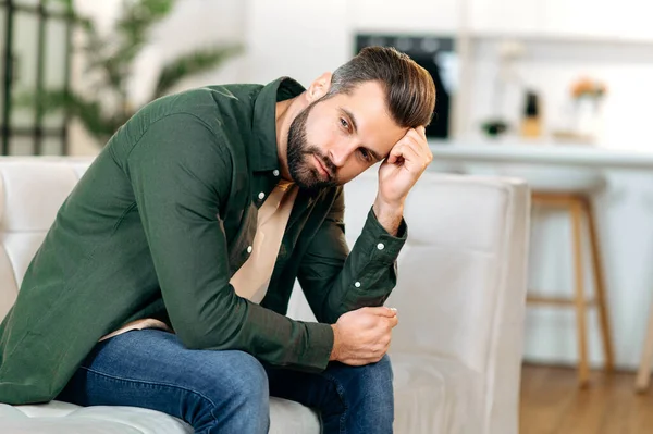 Depression, fatigue, frustration. Upset caucasian man, sit on a sofa in living room, leaning on the hand, experiencing personal problems, stress, loneliness, needs psychological support and rest