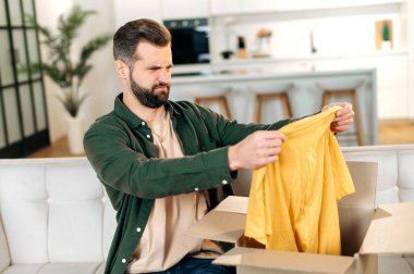 Üzücü beyaz adam, günlük giysiler içinde, oturma odasında bir koltukta oturuyor, paketini açıyor, karton kutudan giysilerini çıkarıyor, hayal kırıklığına uğramış, tatminsiz, haksız beklentilere bakıyor.