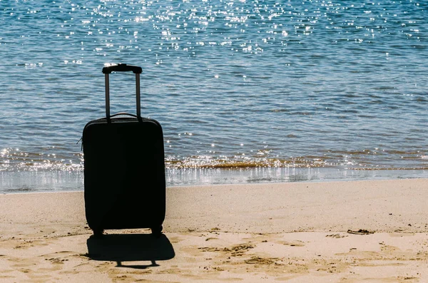 Black travel suitcase on sandy beach with turquoise sea background, summer holidays concept