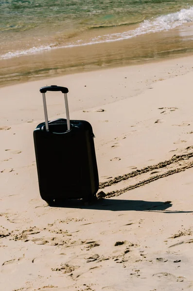 Black travel suitcase on sandy beach with turquoise sea background, summer holidays concept