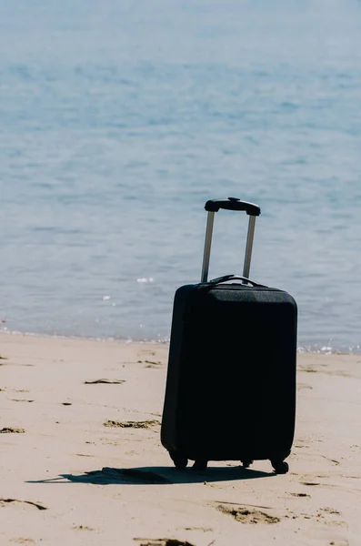 Black travel suitcase on sandy beach with turquoise sea background, summer holidays concept