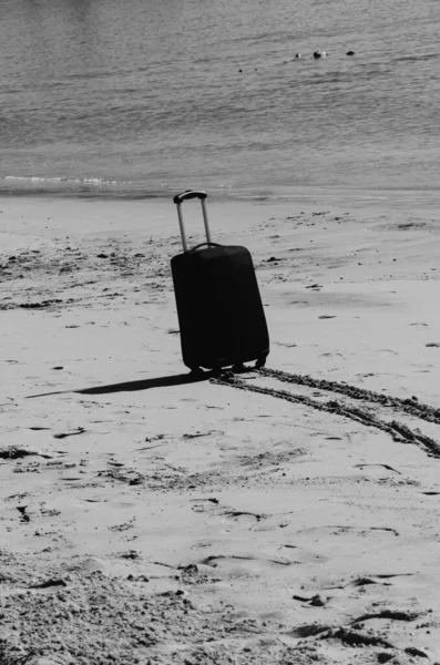 Black travel suitcase on sandy beach with turquoise sea background, summer holidays concept in monochrome