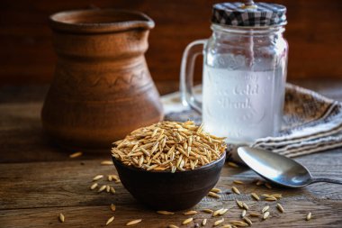 On the wooden kitchen table are a ceramic bowl of oat grains, a glass cup with oat kvass on a kitchen napkin, and an earthen jug. Village life.