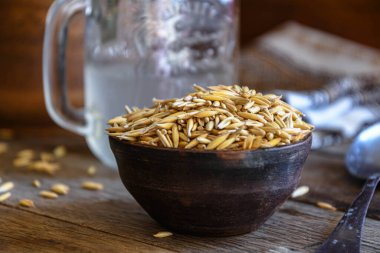 On the wooden kitchen table are a ceramic bowl of oat grains, a glass cup with oat kvass on a kitchen napkin, and an earthen jug. Village life.
