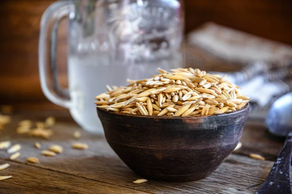 On the wooden kitchen table are a ceramic bowl of oat grains, a glass cup with oat kvass on a kitchen napkin, and an earthen jug. Village life.