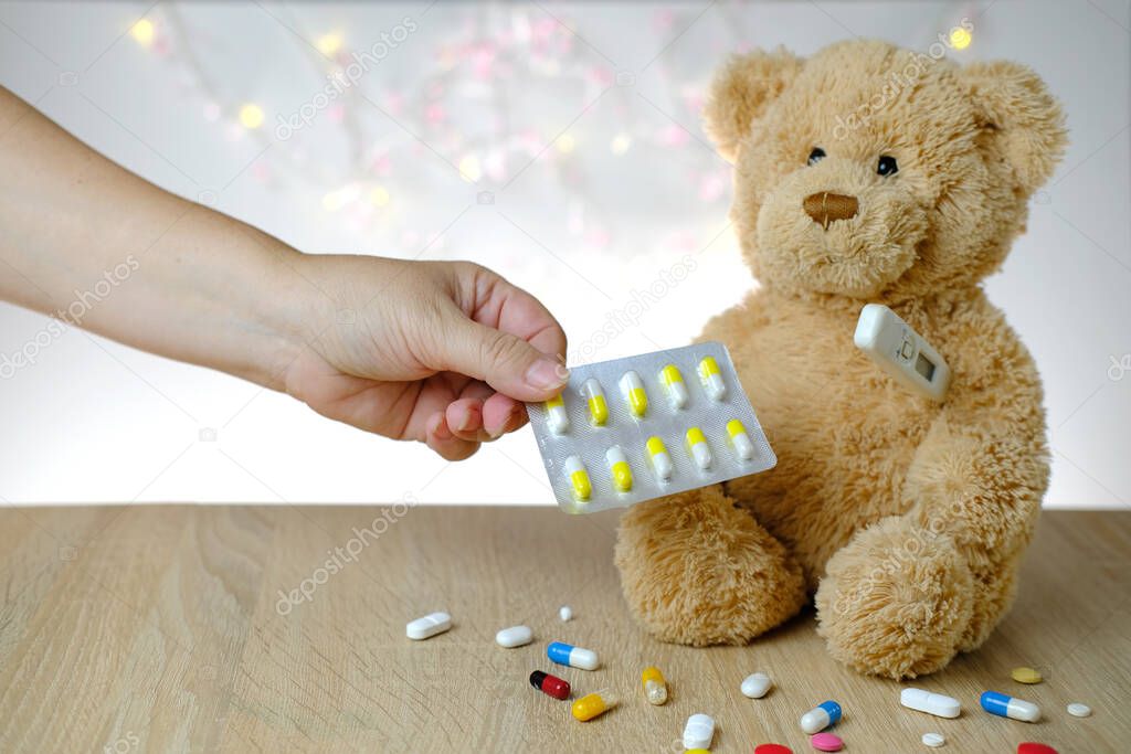close-up of female hand holding blister with pills in hand, teddy bear kids toy with stick thermometer, concept of pediatrics, childhood diseases, treatment and insurance