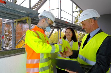 İki adam, bir mimar ve inşaat mühendisi, ustabaşı ve bir kadın, devam etmekte olan çok katlı bir evin inşaat alanında dizüstü bilgisayardaki bir projenin performansını inceliyorlar.