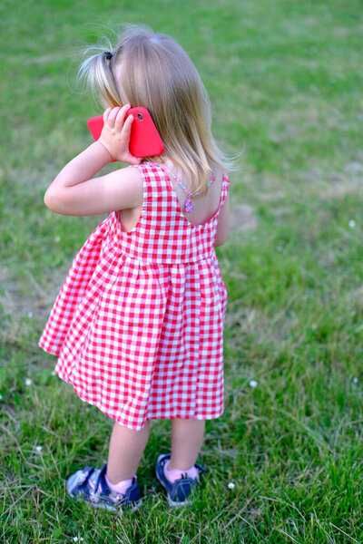 Маленький ребенок, 2-летняя девочка с белыми волосами в красном платье держит смартфон рядом с ухом, общается с друзьями, родителями, концепция детства, ранее умение обращаться с современными технологиями