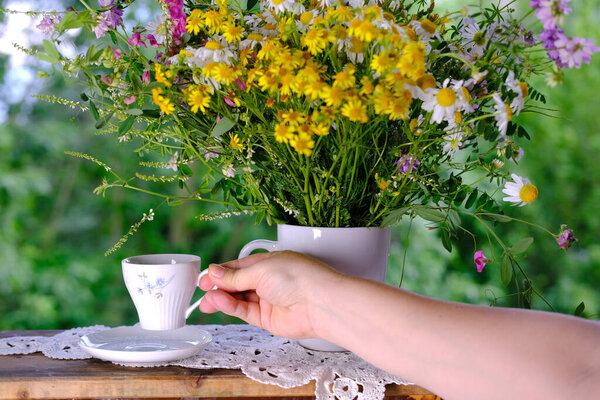 female hands holding a cup of tea, beautiful summer bouquet of wildflowers, handmade lace white doily on the table, summer time concept, tea drinking in the garden