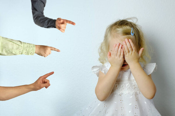 punished small child, blonde girl 2 years old in white dress covered her eyes with her hands, hands of relatives shown with finger, concept of parental abuse, psychological violence, humiliation