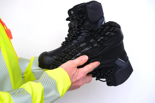 black work boots made of leather with reinforced cape, high top in hands of man, mature male mature builder, worker in yellow work clothes testing special protective professional shoes, for mountains