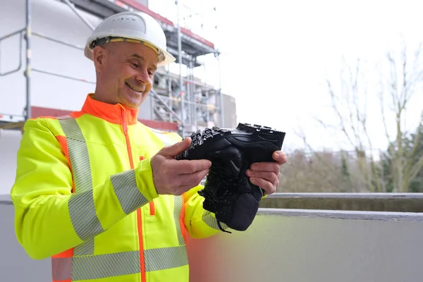 black work boots made of leather with reinforced cape, high top in hands of man, mature male mature builder, worker in yellow work clothes testing special protective professional shoes, for mountains