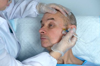 Yetişkin karizmatik bir adamın yakın çekimi, son 60 yıl mavi hastane elbisesi içinde cilt sağlığı ve güzellik kliniğinde yatıyor, doktorun elleri hastaya iğne yapıyor, kozmetik yaşlanmayı önleme prosedürleri konsepti