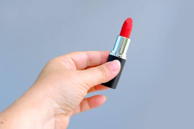 Gri arka planda parlak kırmızı ruj izi olan bir tüp tutan yakın plan kadın eli makyaj yapmak istiyor, dekoratif kozmetik konsepti, kadın günlük rutini.