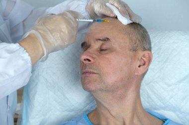 Yetişkin karizmatik bir adamın yakın çekimi, son 60 yıl mavi hastane elbisesi içinde cilt sağlığı ve güzellik kliniğinde yatıyor, doktorun elleri hastaya iğne yapıyor, kozmetik yaşlanmayı önleme prosedürleri konsepti