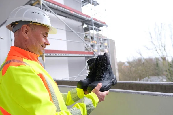 black work boots made of leather with reinforced cape, high top in hands of man, mature male mature builder, worker in yellow work clothes testing special protective professional shoes, for mountains