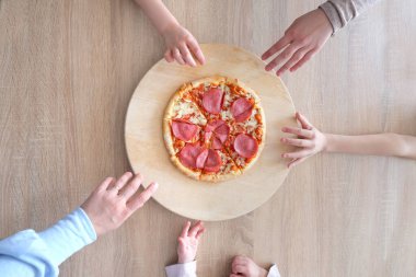 Masanın üzerinde yuvarlak bir tepside salamlı sıcak iştah açıcı bir pizza yatıyor, büyük bir aile masada oturuyor, çocukların elleri pizza dilimlerini alıyor, ev yemeği konseptini, çocukların lezzet etkilerini alıyor.