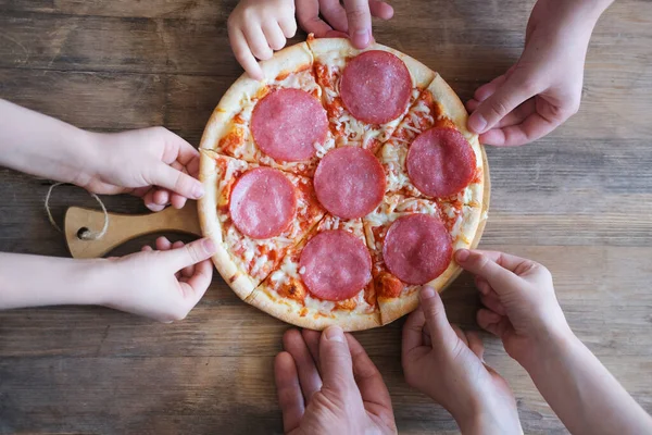 Masanın üzerinde yuvarlak bir tepside salamlı sıcak iştah açıcı bir pizza yatıyor, büyük bir aile masada oturuyor, çocukların elleri pizza dilimlerini alıyor, ev yemeği konseptini, çocukların lezzet etkilerini alıyor.