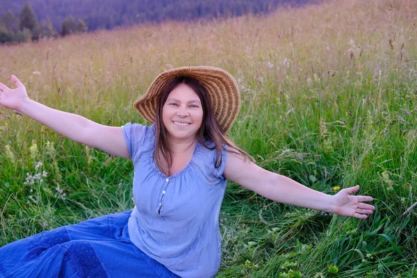 成熟した女性45歳ウィッカー帽子と青い服で山の緩やかな斜面に座って 喜んで彼女の腕 緑の草 自然上のピクニックの概念を広げ 人生と自然 アクティブなライフスタイルをお楽しみください — ストック写真