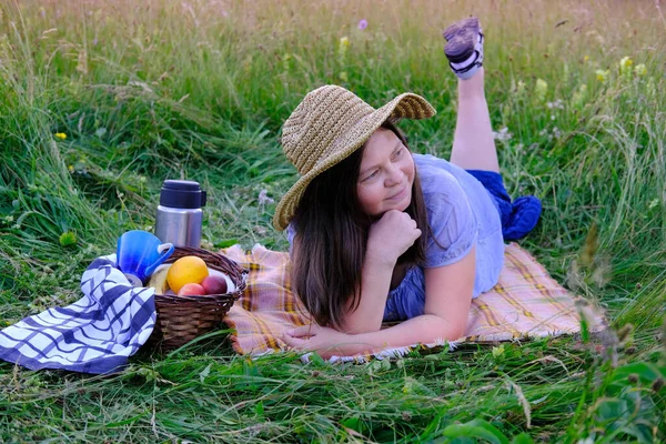 woman 45-50 years old in wicker hat and blue clothes lies on grass in meadow, next to picnic basket with fruit, thermos, concept of family picnic on nature, enjoy life and nature, active lifestyle