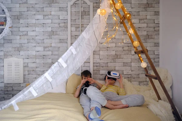 two children 8-10 years old, boys lying on bed in tent in modern VR glasses interacting with network while having virtual reality experience, study of another world, modern teaching technologies