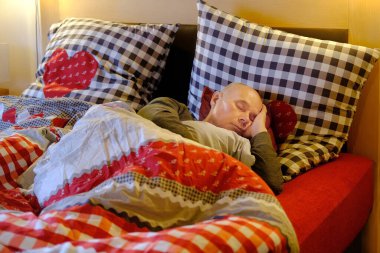 60 'larında olgun bir adam yatakta, otel odasında yatakta, battaniye, iki yastık, kırmızı pamuklu çarşaf, sağlıklı uyku konsepti, uykusuzluk problemleri, otelde rahat bir konaklama.