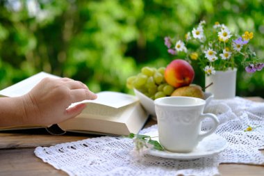 çay, fincan tabağında beyaz bardak kahve, açık kitap, meyve, dantel peçete, bahçedeki ahşap masa üzerinde yabani çiçekler, yeşil yapraklı bulanık doğal arka plan, kavram çay saati