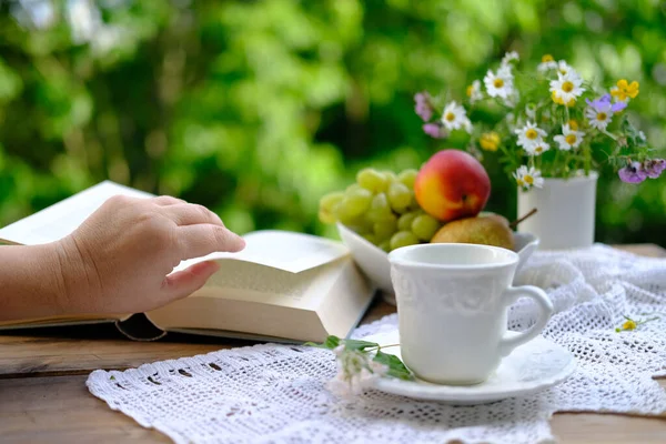 çay, fincan tabağında beyaz bardak kahve, açık kitap, meyve, dantel peçete, bahçedeki ahşap masa üzerinde yabani çiçekler, yeşil yapraklı bulanık doğal arka plan, kavram çay saati