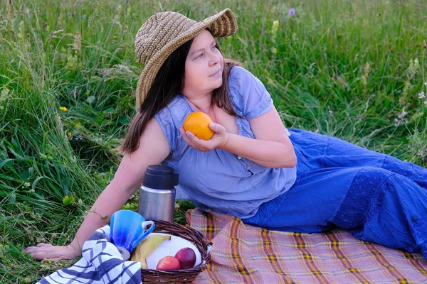 woman 45-50 years old in wicker hat and blue clothes sits on grass in meadow, orange in her hand, next to picnic basket with fruit, thermos, concept of family picnic on nature, enjoy life and nature