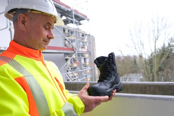 black work boots made of leather with reinforced cape, high top in hands of man, mature male mature builder, worker in yellow work clothes testing special protective professional shoes, for mountains