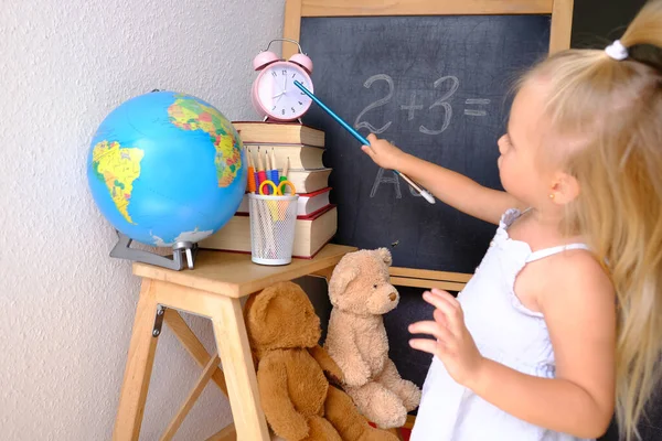 child, blonde girl plays school, small teacher teaches lesson, student home office, alarm clock, books, teddy bears, chalk board, globe, concept of education, back to school, knowledge day