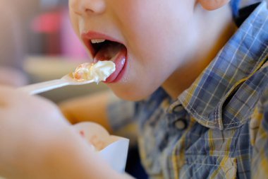 cold ice cream with fruit jam on wooden spoon close-up, child 7 years old, boy appetizing eating strawberry dessert, happy childhood concept, children's delicacy, healthy and unhealthy food