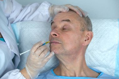 Yetişkin karizmatik bir adamın yakın çekimi, son 60 yıl mavi hastane elbisesi içinde cilt sağlığı ve güzellik kliniğinde yatıyor, doktorun elleri hastaya iğne yapıyor, kozmetik yaşlanmayı önleme prosedürleri konsepti