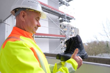 Deriden yapılmış, güçlendirilmiş pelerinli siyah iş botları insan eli altında, olgun erkek müteahhit, sarı iş elbiseleri giyen işçi özel koruyucu profesyonel ayakkabıları test ediyor, dağlar için.