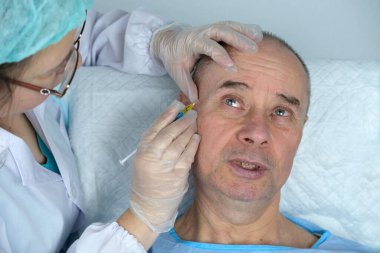 Yetişkin karizmatik bir adamın yakın çekimi, son 60 yıl mavi hastane elbisesi içinde cilt sağlığı ve güzellik kliniğinde yatıyor, doktorun elleri hastaya iğne yapıyor, kozmetik yaşlanmayı önleme prosedürleri konsepti
