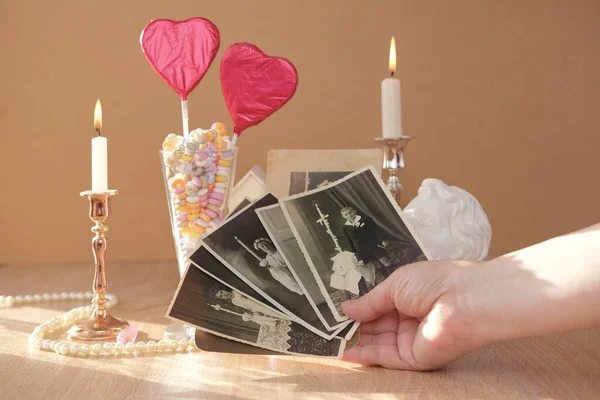 stack of vintage photos in female hand, romantic still life in love style, in glass vase red hearts on sticks, colorful candies, candles burning, concept of family tree, genealogy, childhood memories