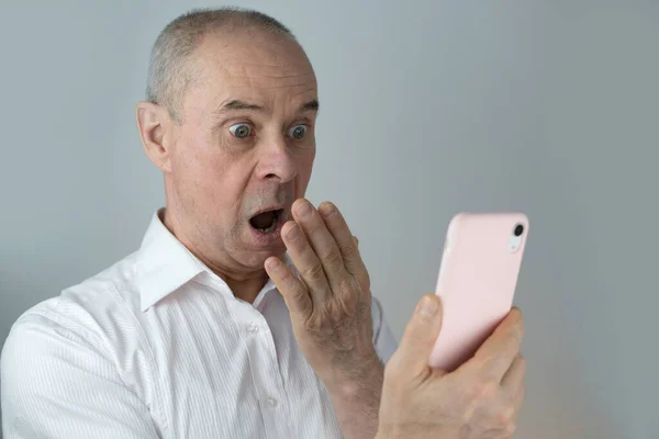 成熟而有魅力的男人的特写看着粉红的手机屏风 六十岁高龄的老人惊讶不已 被新闻 明亮的面部表情 概念等吓到了一大跳 — 图库照片