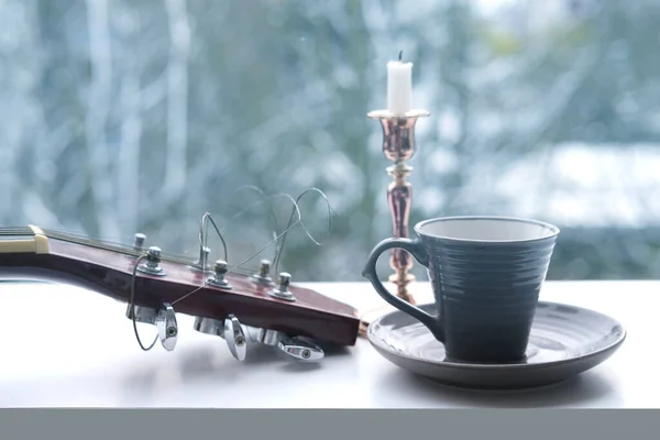 guitar with strings, a cup of coffee, tea, a candle in a candlestick against the backdrop of a beautiful winter landscape