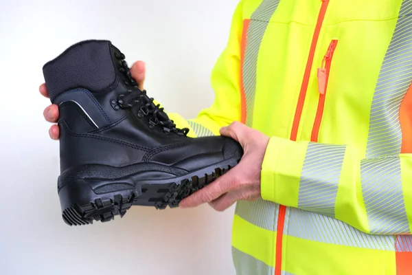 black work boots made of leather with reinforced cape, high top in hands of man, mature male mature builder, worker in yellow work clothes testing special protective professional shoes, for mountains