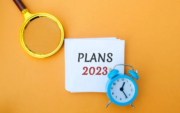 Note with words Plans 2023. Setting goals and planning targets for the next year. Motivation, vision. Business and finance concept. Magnifying glass and alarm clock