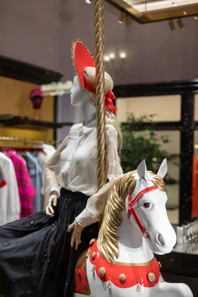 Escaparate decorado festivamente de una tienda de mujeres. Un maniquí en un sombrero rojo se sienta en un caballo de Navidad de juguete — Foto de Stock