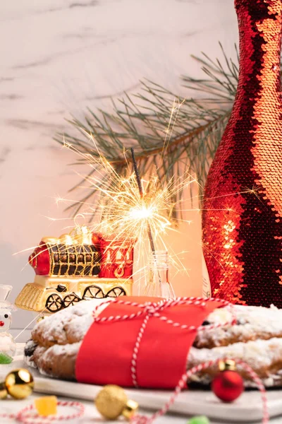 Festive Christmas concept. Homemade cake stollen , Christmas toys, a burning sparkler and a bottle of wine — Stock Photo, Image