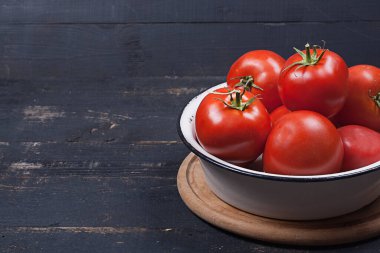 A lot of red tomatoes in an iron white plate on a black wooden table close-up