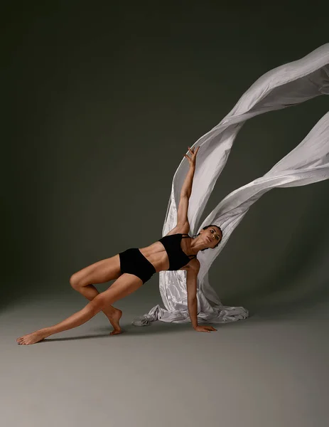 Эмоциональная танцовщица балета в костюме и летающем материале позирует в танце в различных позах на сером фоне — стоковое фото