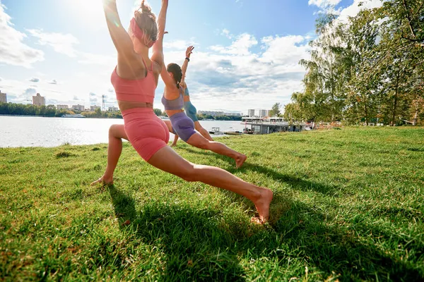 Drei fitte schöne Frauen beim Virabhadrasana-Training im Park an einem Sommertag, in Sportkleidung, in voller Länge. Lifestyle-Konzept. — Stockfoto