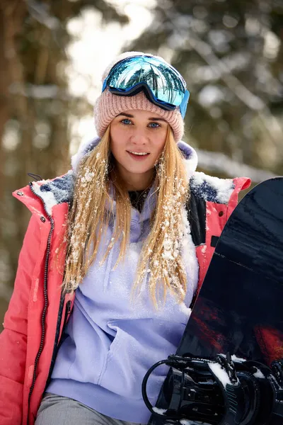 Kaukasische Snowboarderin hält Snowboard auf wunderschönem verschneiten Waldhintergrund bei sonnigem Tag. — Stockfoto