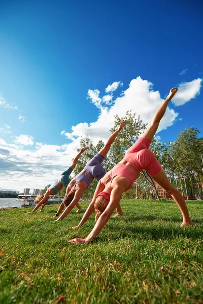 Tres hermosas mujeres en forma haciendo Virabhadrasana, haciendo ejercicio en el parque en el día de verano, usando tops de ropa deportiva, de cuerpo entero. Concepto de estilo de vida. — Foto de Stock