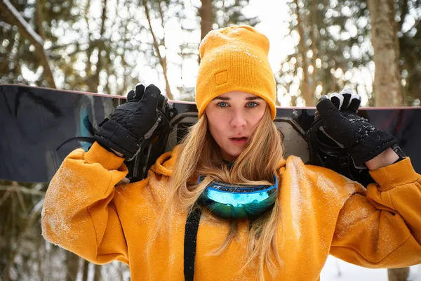 Niña o mujer snowboarder entra en los deportes de invierno en el bosque nevado que se para en la nieve y sostiene una tabla de snowboard —  Fotos de Stock