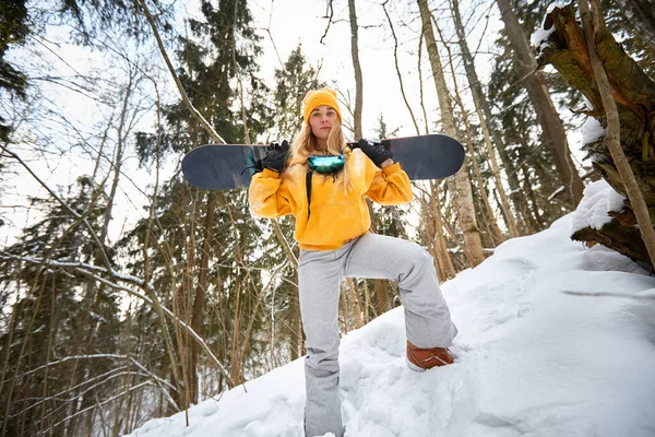 Młoda dziewczyna lub kobieta snowboardzista idzie na sporty zimowe w zaśnieżonym lesie stoi w śniegu i trzyma snowboard — Zdjęcie stockowe