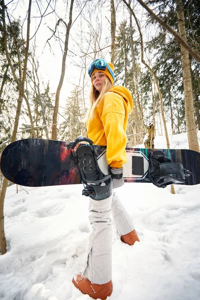 Niña o mujer snowboarder entra en los deportes de invierno en el bosque nevado que se para en la nieve y sostiene una tabla de snowboard —  Fotos de Stock